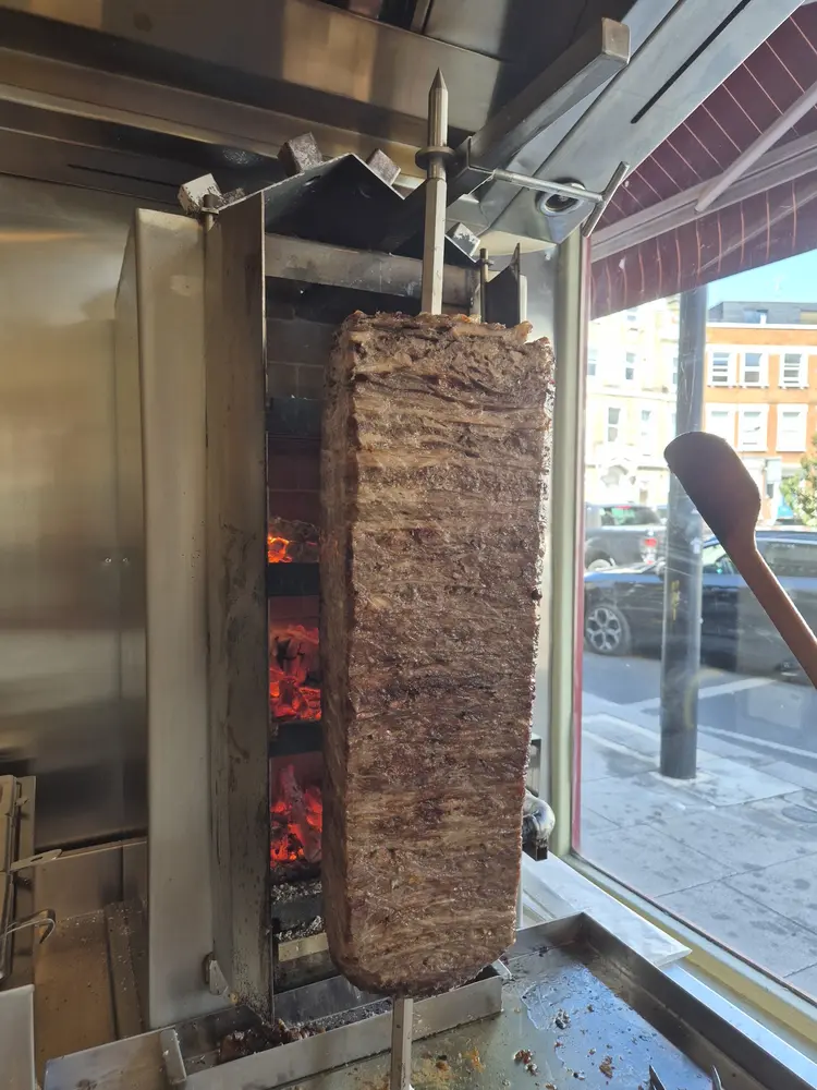 Tall veal döner spit cooking beside glowing wood embers in the window